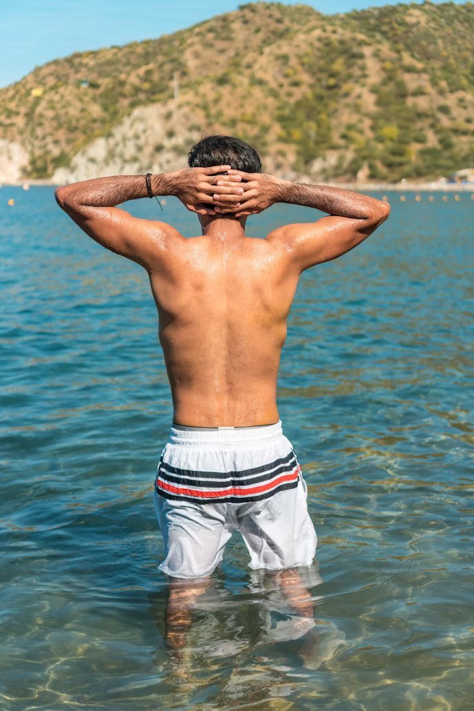 Before: Before and after AI preview of rounder, fuller shoulders and rear delts on a man standing in the sea with hands on his head, made in Buldy